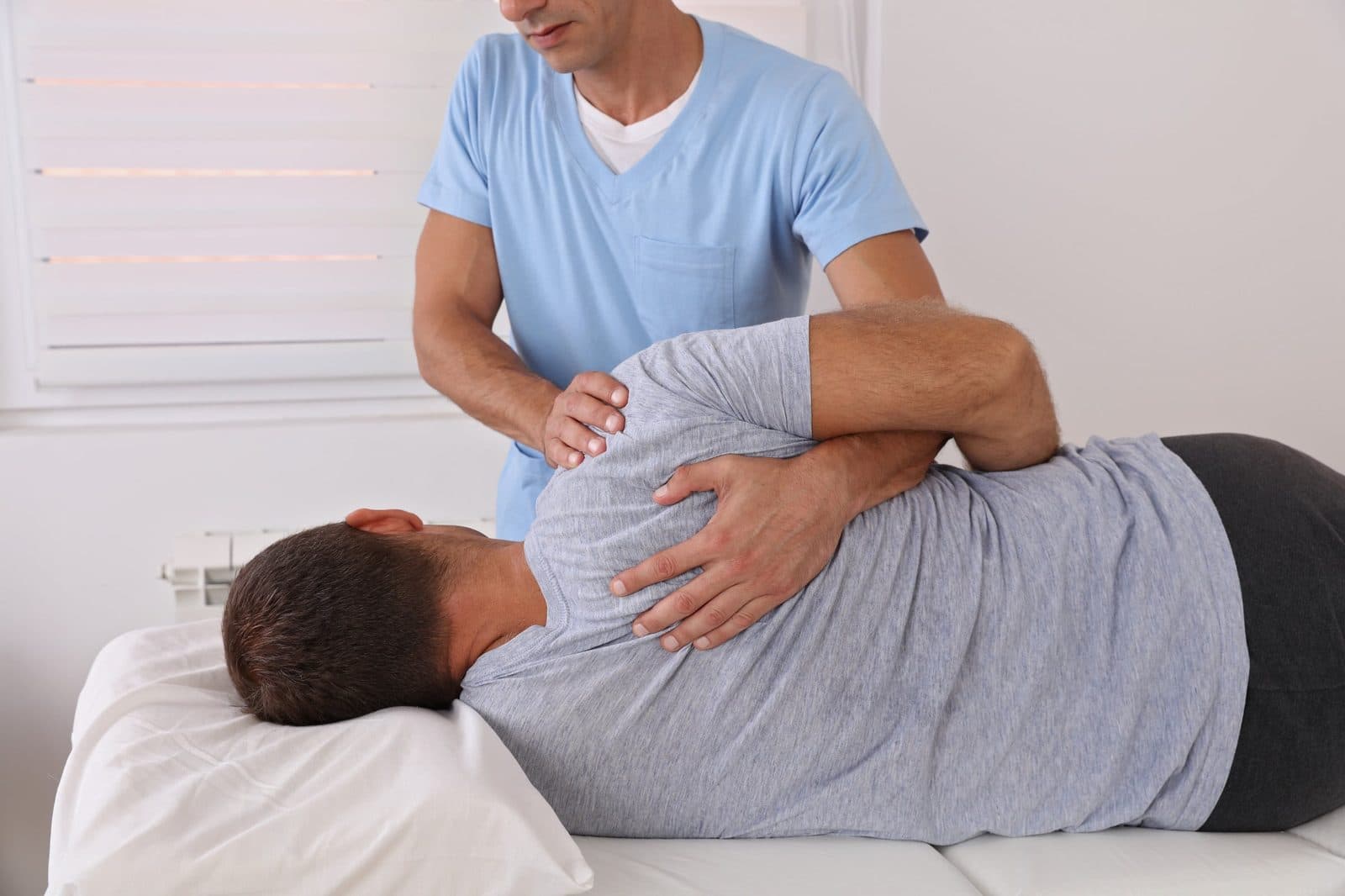 Manual therapy on a patient lying on a treatment table in a bright room with window blinds.